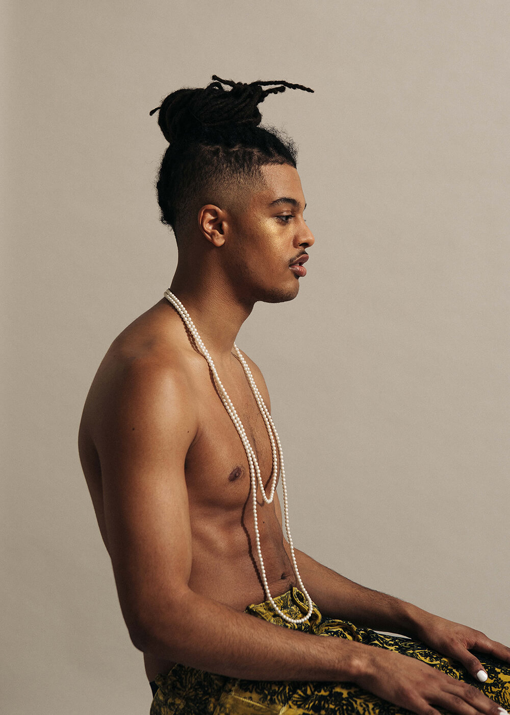A editorial studio portrait of a shirtless man sitting wearing beads