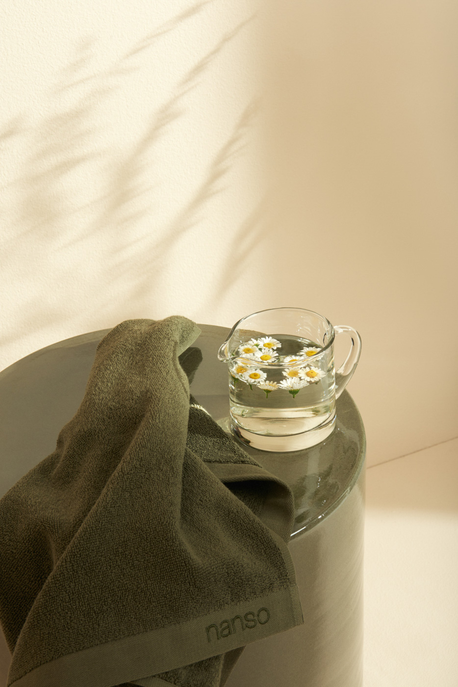 Green_towel_and_glass_jug_containing_daisies_and_water_on_ceramic_stool_on_beige_background_in_sunlight_for_Nanso_Home_collection_photographed_by_Sara_Lehtomaa