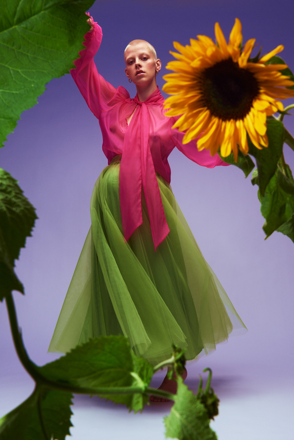 Pregnant_short_haired_blond_woman_wearing_pink_blouse_and_green_tulle_skirt_behind_sunflowers_on_lilac_background_photographed_by_Sara_Lehtomaa