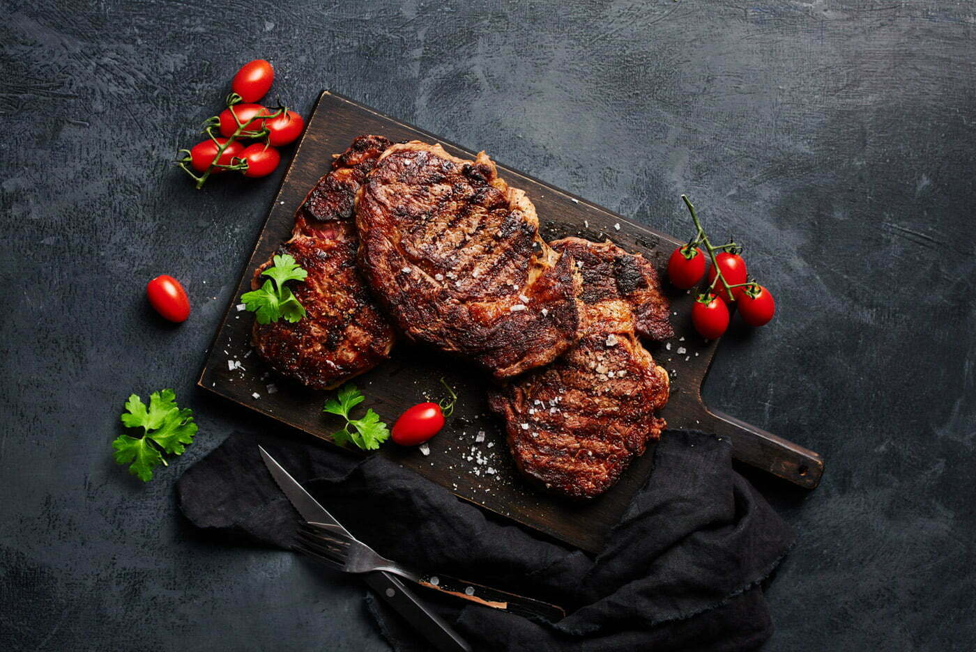 three_steaks_and_cherry_tomatoes_on_a_cutting_board_with_a_dark_gray_background_by_Elina_Himanen