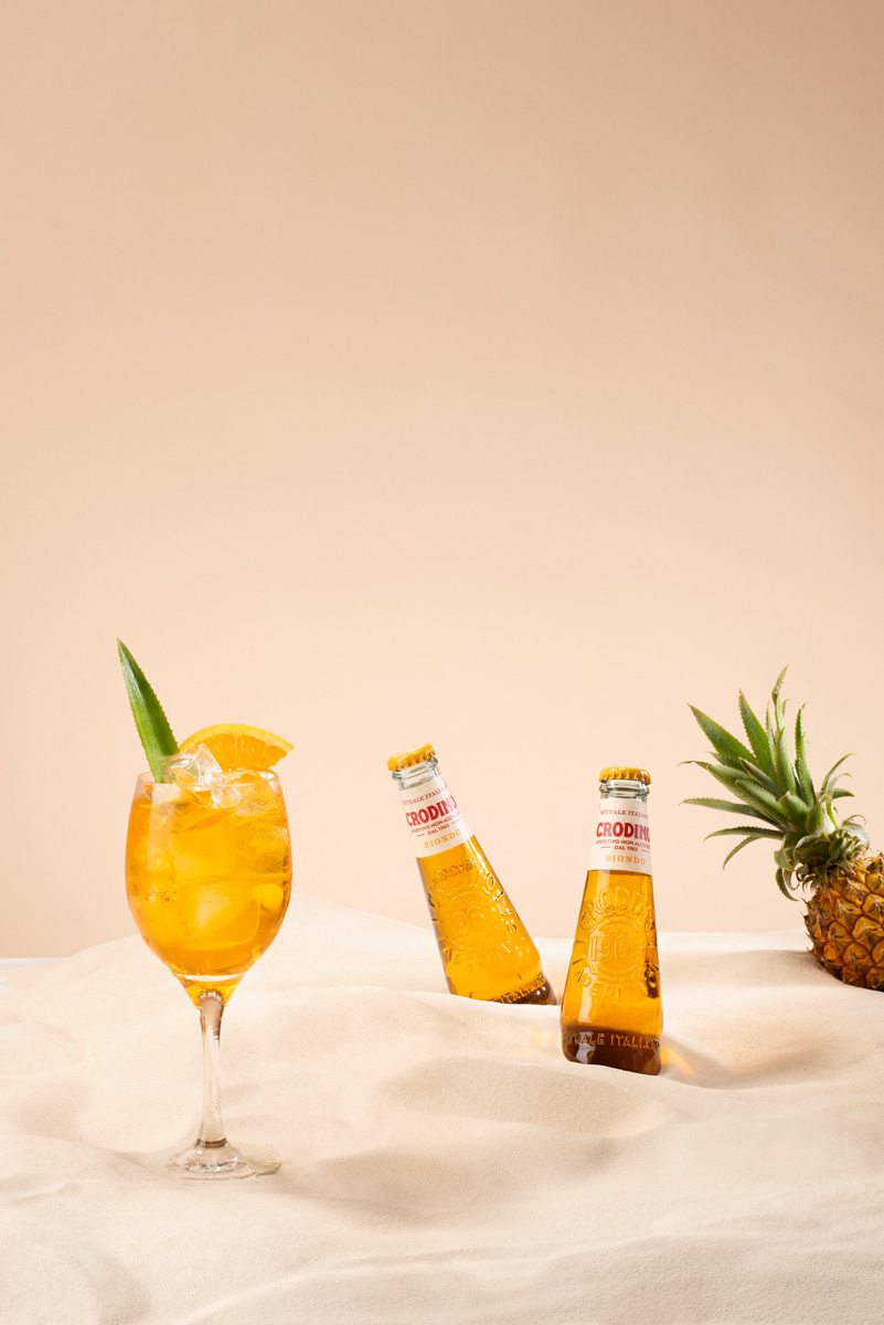 two crodino bottles and a glass placed in sand with a pineapple by Tomas Olsen
