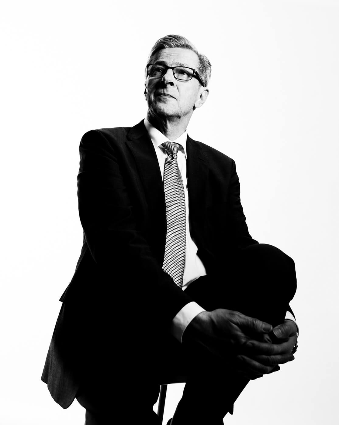 A black and white corporate portrait of a man wearing a suit sitting on a chair
