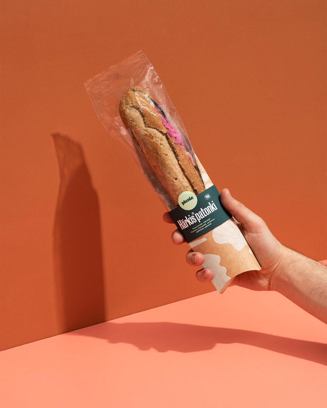 a hand holding a baguette wrapped in fresh package on a orange and pink background by Tomas Olsen